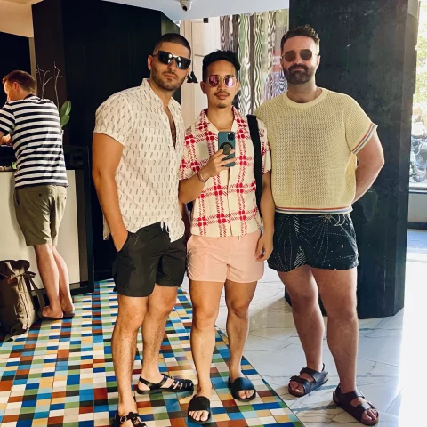 Three men dressed in tshirts and shorts, wearing sunglasses, standing on a colorful floor.