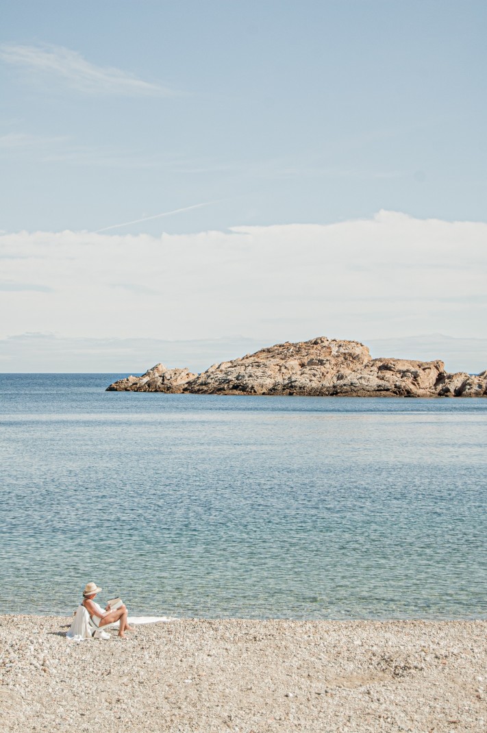 A woman on a pebble beach in Costa Brava, Spain with crystal blue waters and a rocky formation.