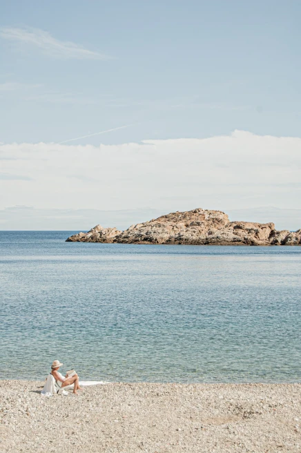 A woman on a pebble beach in Costa Brava, Spain with crystal blue waters and a rocky formation.