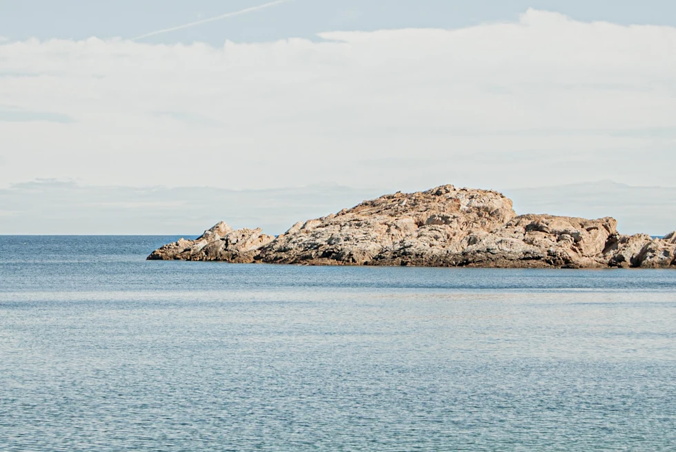 A woman on a pebble beach in Costa Brava, Spain with crystal blue waters and a rocky formation.