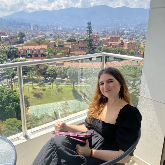 travel advisor Laura Zapata wears a black dress and sits on a balcony overlooking a green city and mountains in the distance