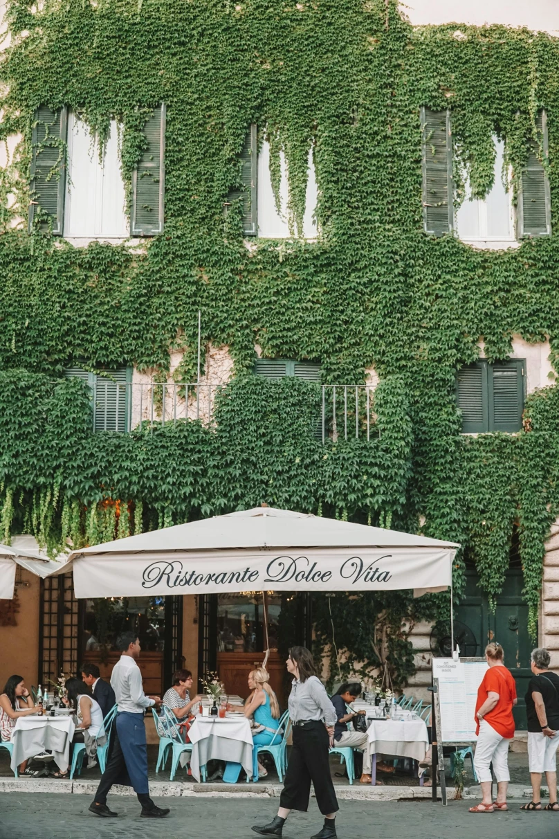 People dining at outdoor tables of a Roman restaurant, covered in green vines.