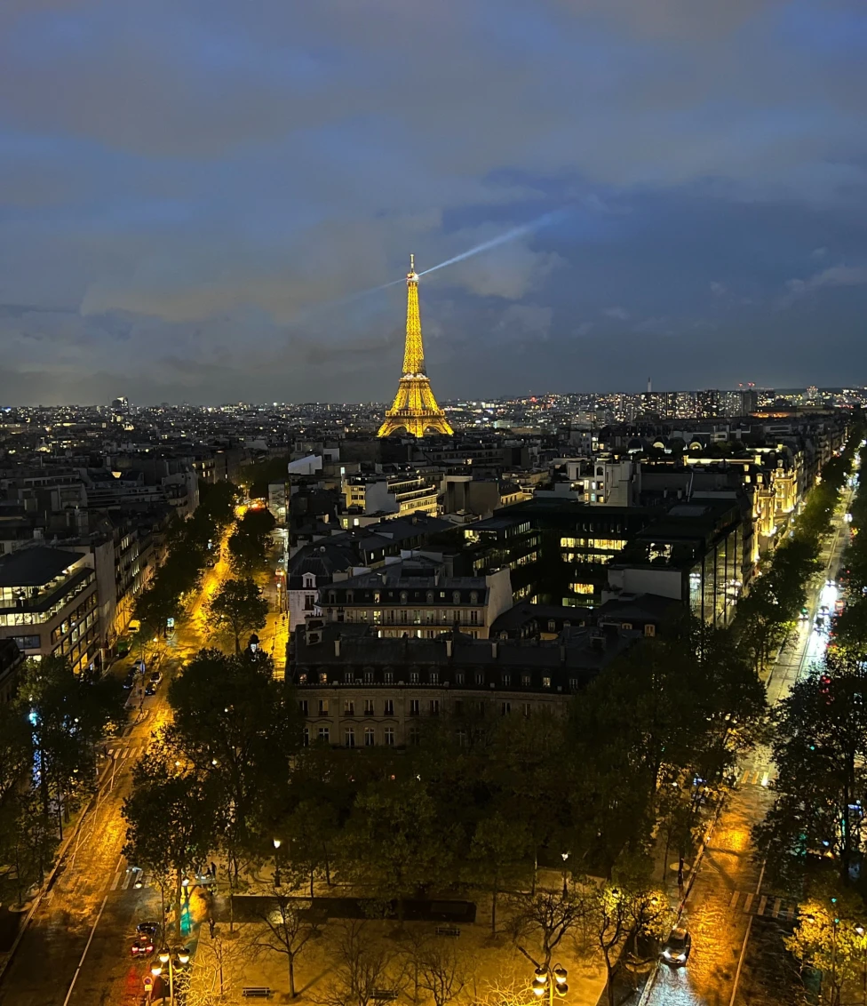 A view from a Parisian rooftop with the city lit up below at night, the Eiffel Tower shining with lights in the distance.
