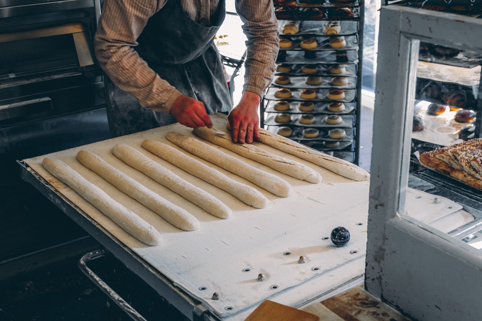 A baker in Iceland working on seven white dough baguettes with red gloves and a blue apron.
