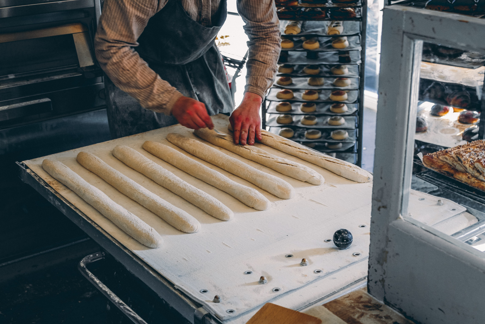 A baker in Iceland working on seven white dough baguettes with red gloves and a blue apron.