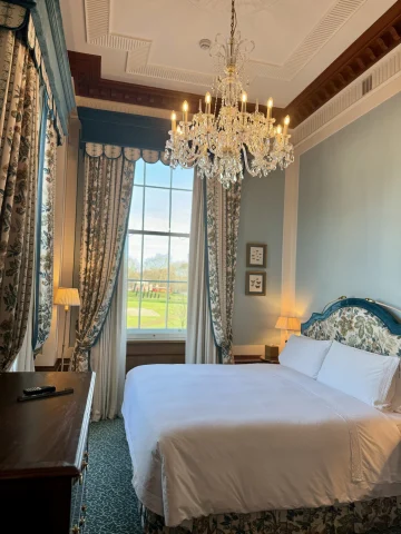 A picture of a hotel suite, complete with a white bed, blue-toned and patterned headboard, crystal chandelier, wooden table, lamps that are turned on, patterned curtains and blue walls.