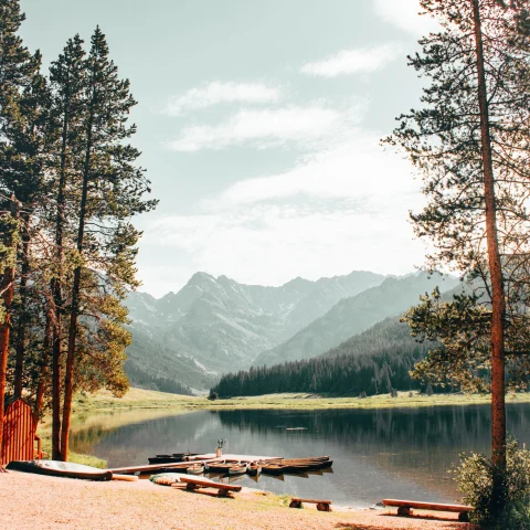 a calm lake with docked canoes at the base of a woodsy mountain region with tall trees framing the scene