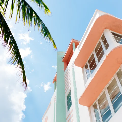 View of coral and white building next to a palm tree with a blue sky