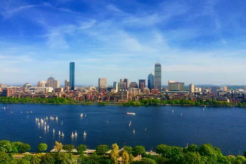 A beautiful view of the Boston skyline with the vibrant blue harbor in the forefront. There are boats and trees surrounding the skyscrapers, water and industrial views. 