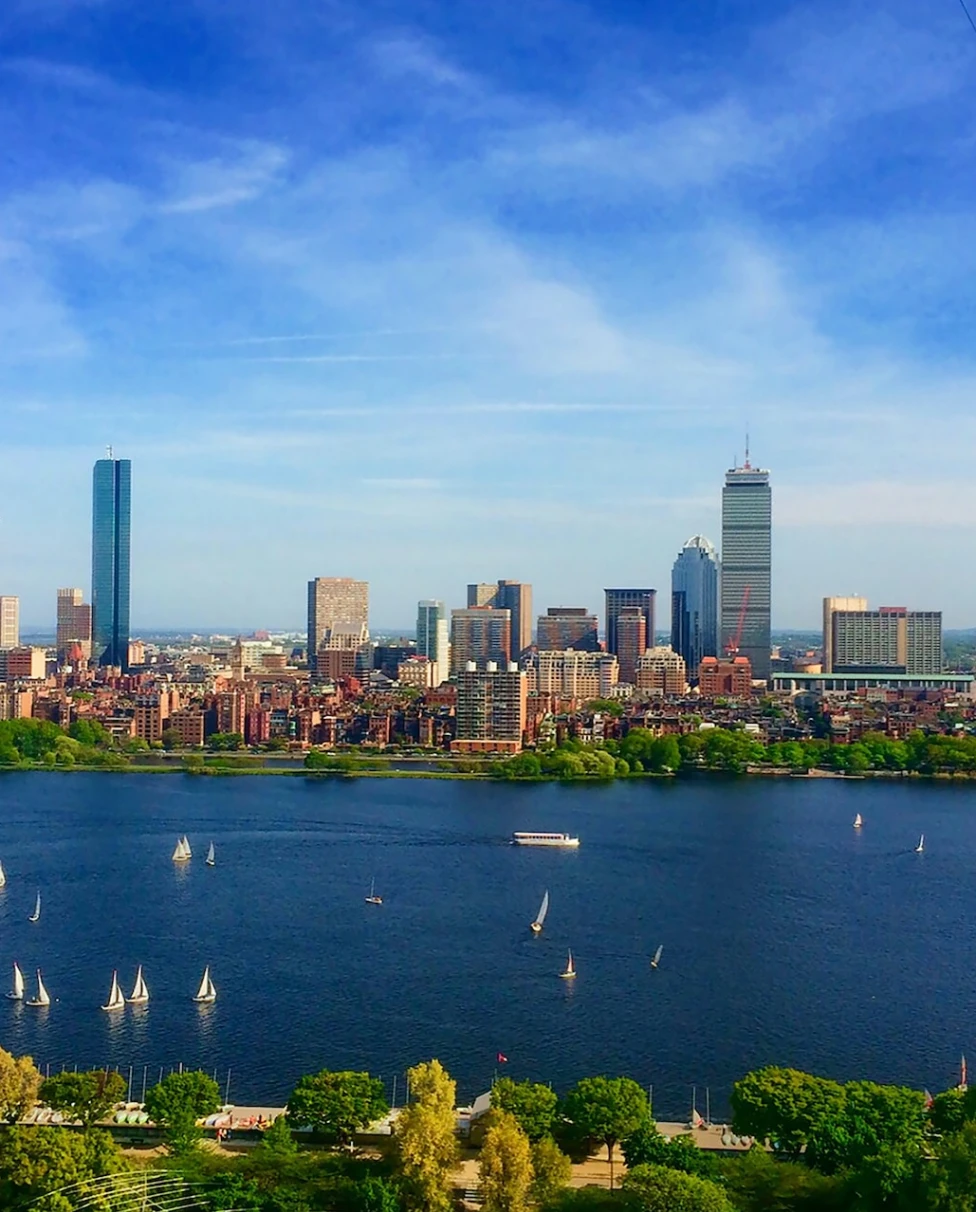 A beautiful view of the Boston skyline with the vibrant blue harbor in the forefront. There are boats and trees surrounding the skyscrapers, water and industrial views. 