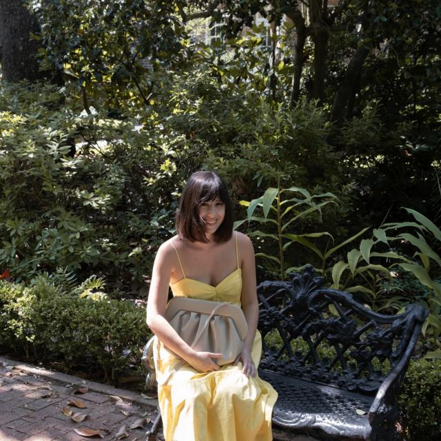 Woman wearing yellow dress sitting on black bench with green foliage in background