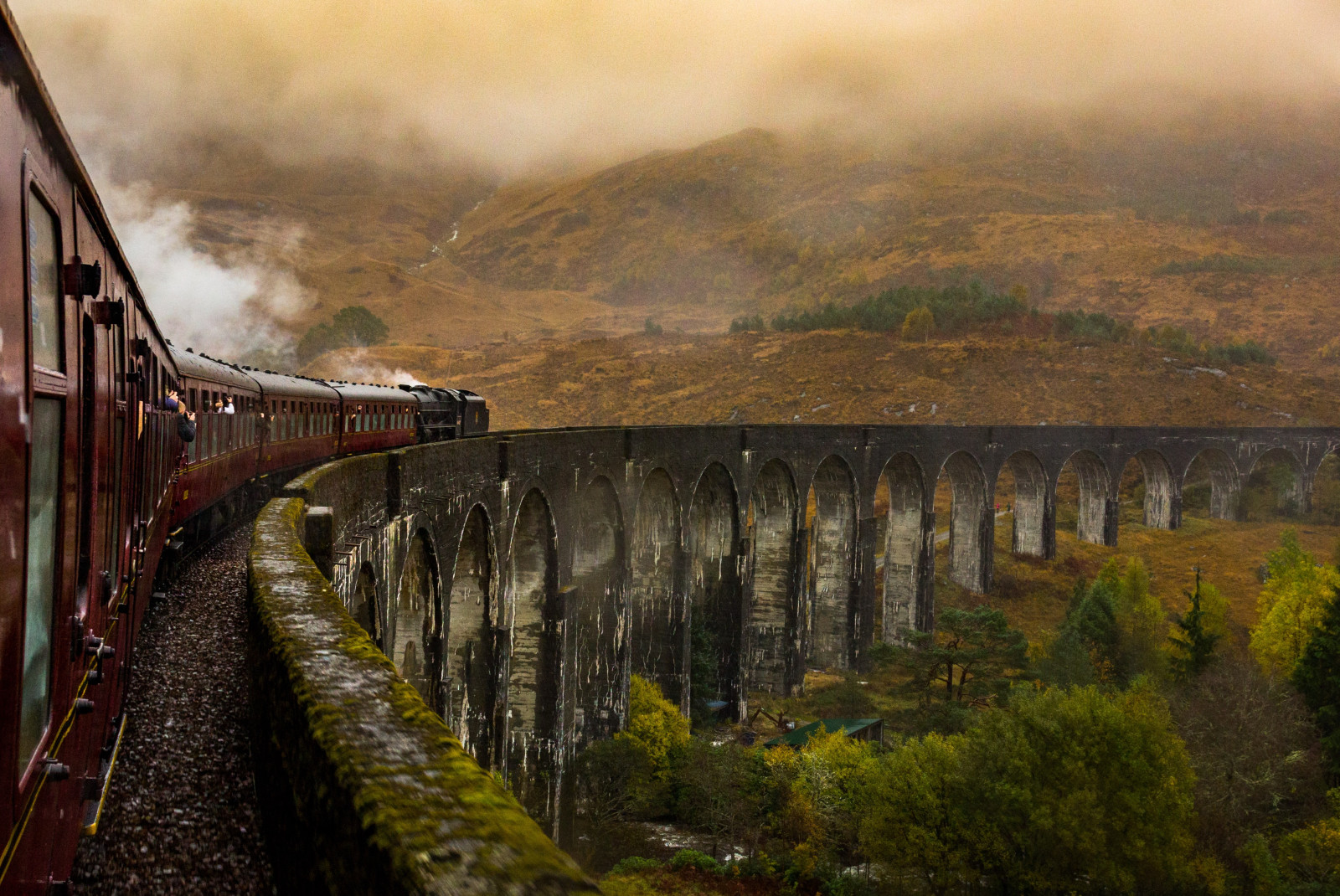 The Jacobite Express in Scotland, a long red train with clear windows steaming ahead over a gray stone bridge with arches and green moss, brown grassy hills and tall green trees below and in the distance with misty clouds.