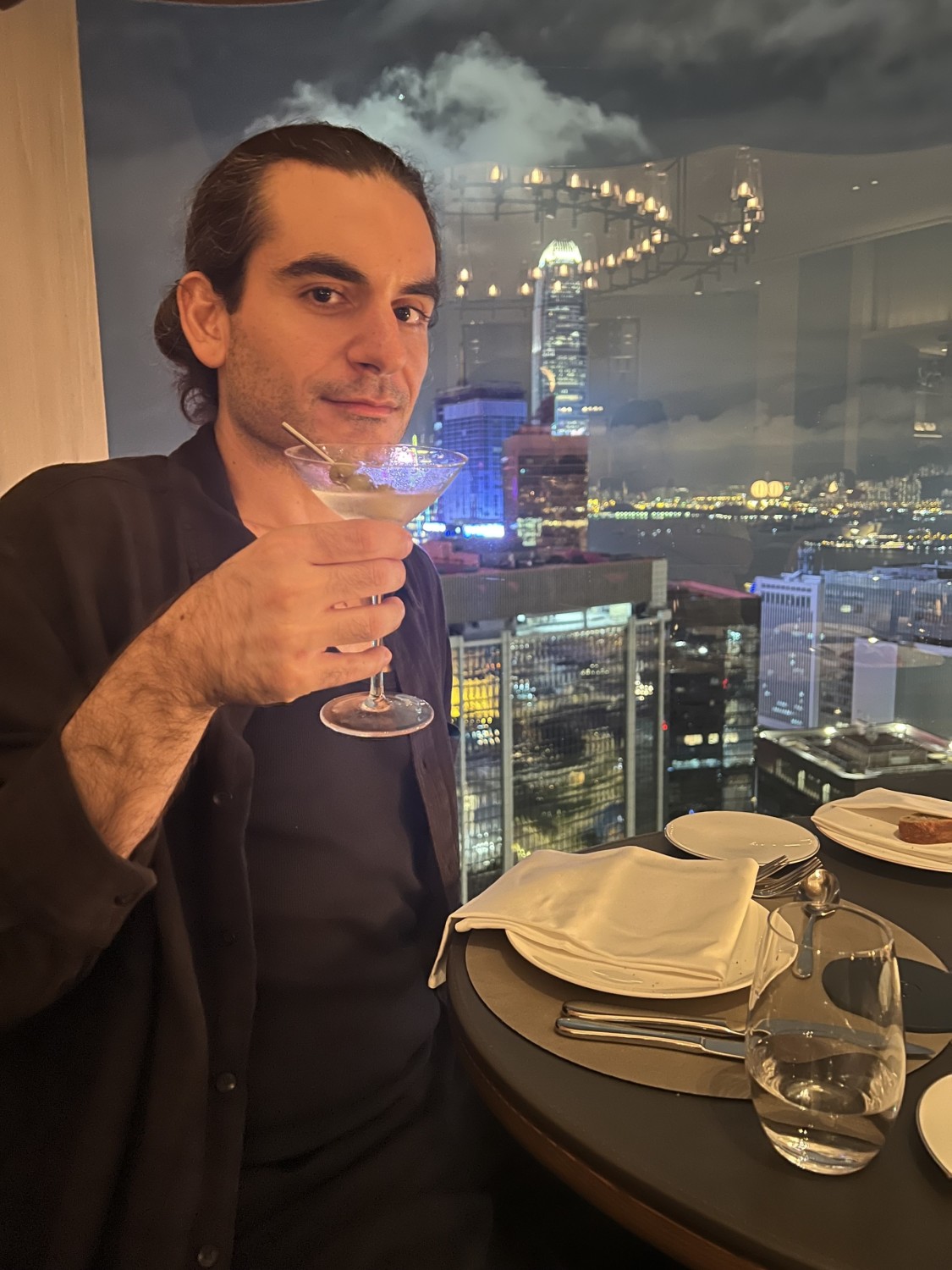 Alex raising his cocktail, at a table of the hotel's bar with a view of Hong Kong in the background.