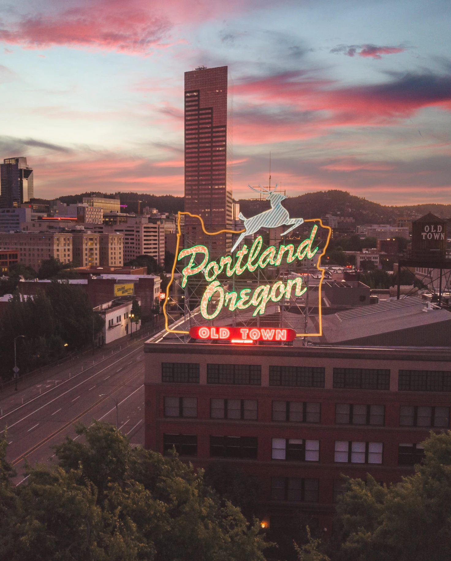 A city view of Portland, Oregon at sunset with pink clouds and a neon sign reading 'Portland Oregon Old Town' with a white deer. 