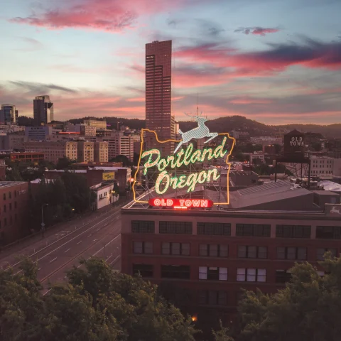 A city view of Portland, Oregon at sunset with pink clouds and a neon sign reading 'Portland Oregon Old Town' with a white deer. 