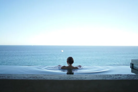 Woman relaxing in infinity pool overlooking the ocean on a bright day.