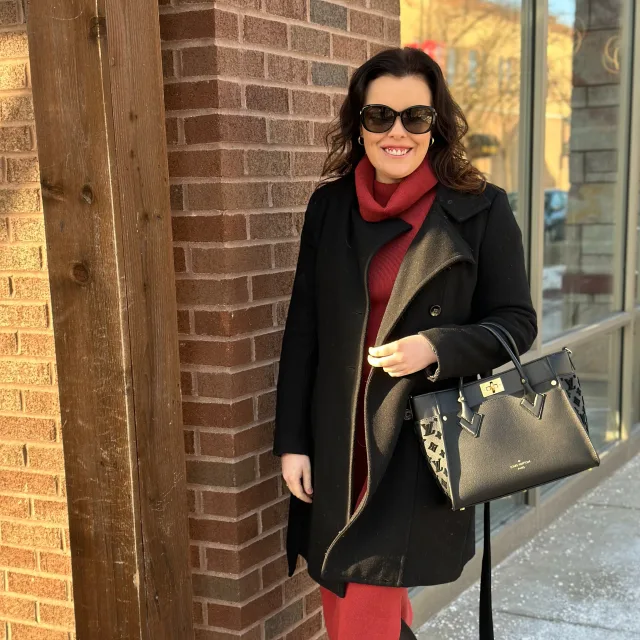 Travel Advisor Riley Wharton in a red turtleneck dress, sunglasses and a black coat in front of a brick wall.