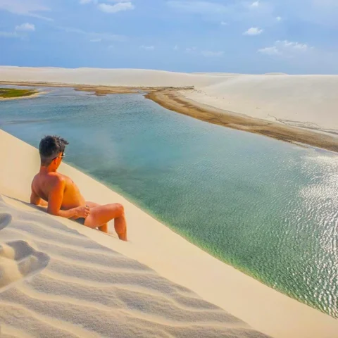 The image depicts a person enjoying a serene view of a calm water body from the edge of a white sand dune.