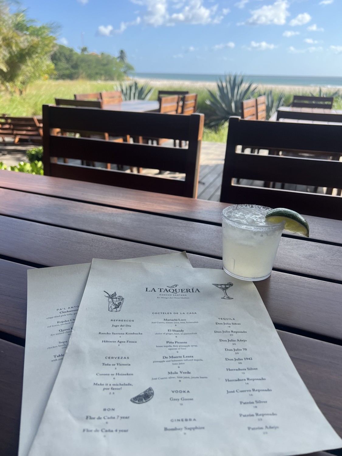 Wooden table at an outdoor restaurant with a menu and cocktail, overlooking a beautiful view