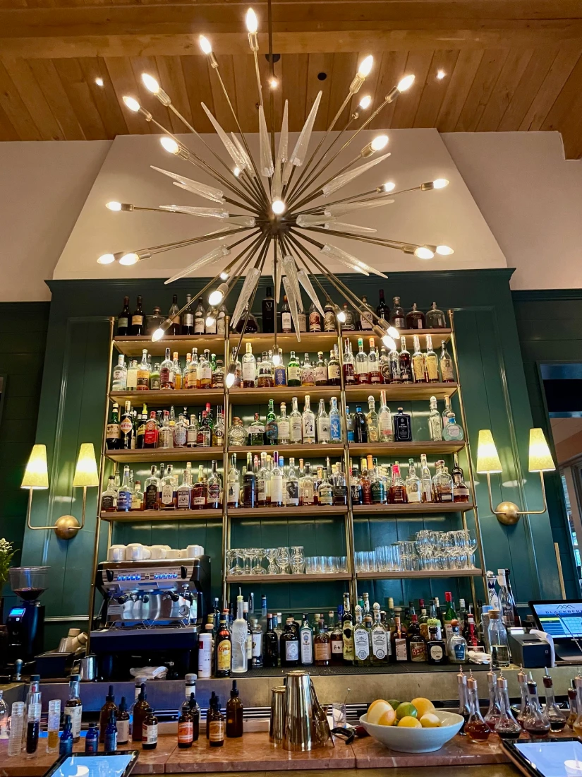 A restaurant bar with a large mid-century modern chandelier, green walls, sconces and shelves full of liquor. 