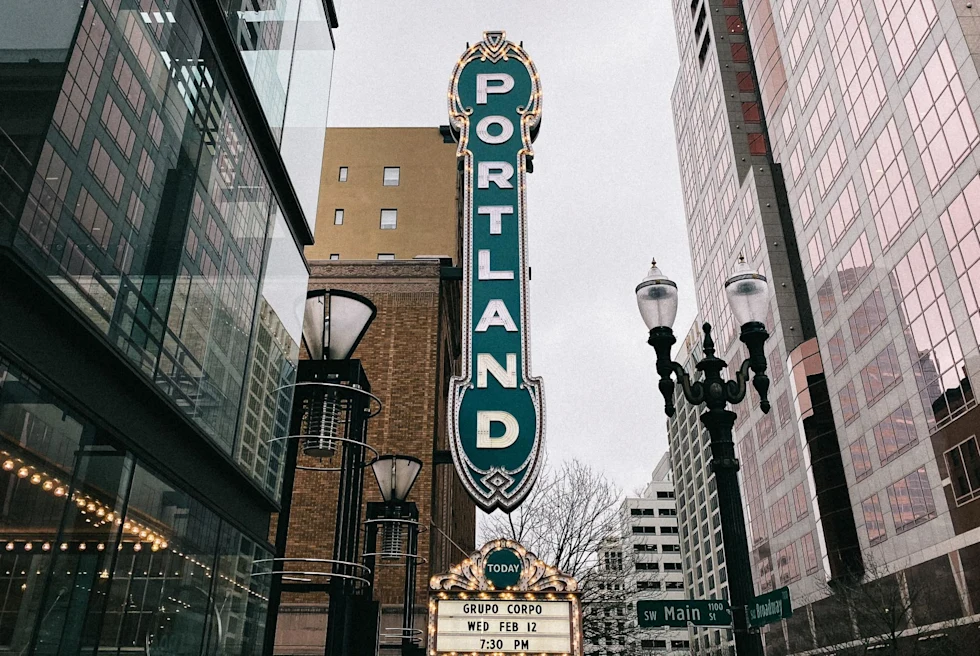 long old school vertical sign over a theater reads, "portland" in white letters, surrounded by large glass high rise buildings