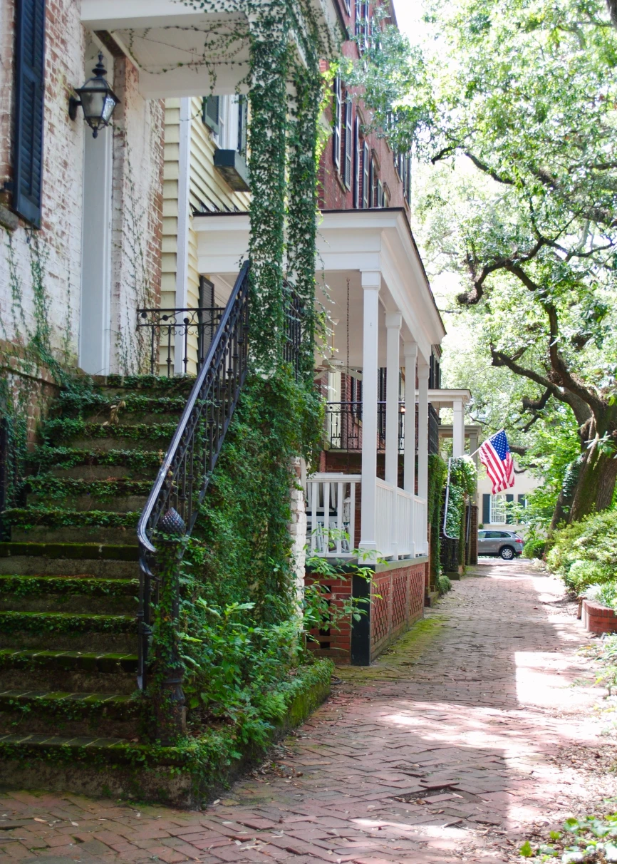 Black staircase covered in green ivy with a brick sidewalk surrounded by green trees with American flag in the distance.