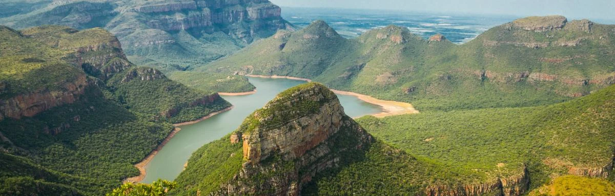 Mountains and rock formations surrounded by greenery and vegetation next to large river in Blyde River Canyon, South Africa.