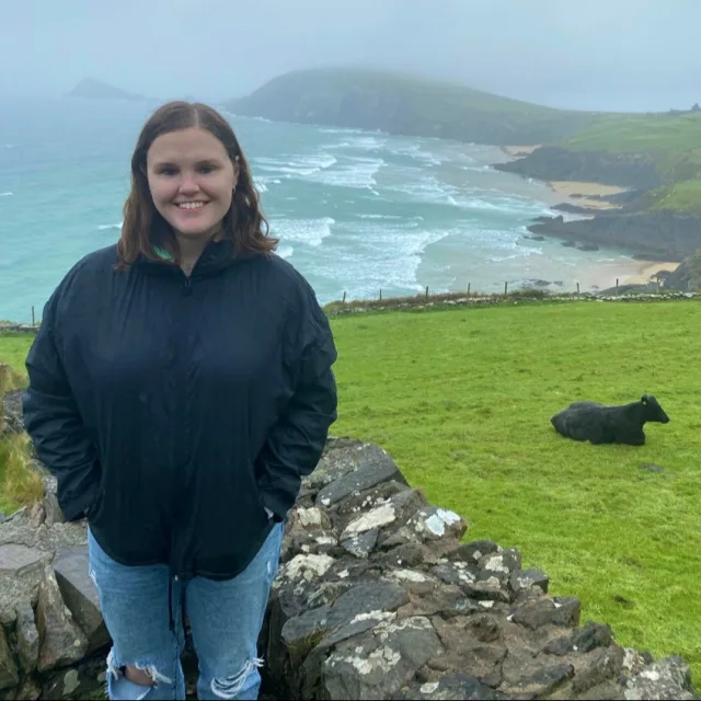 Paige wearing a black top against a green field with a black cow seated in the grass overlooking the sea
