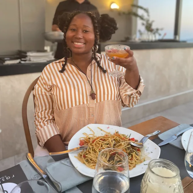 Jia Barclay wearing an orange and white striped top, holding a drink and eating a bowl of pasta 