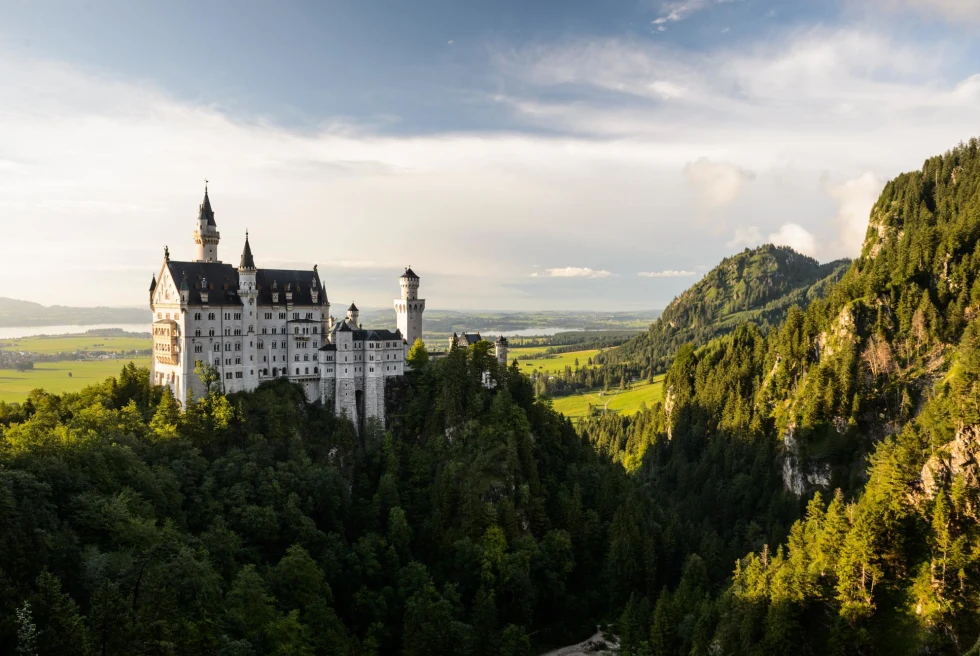 Neuschwanstein Castle on grassy hilltop and alongside lush mountains in Germany.