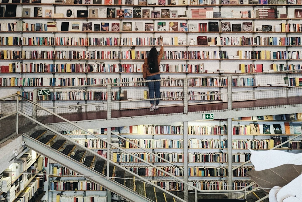 a woman picks a book in a library wall covered in books with a stairwell to a lower floor