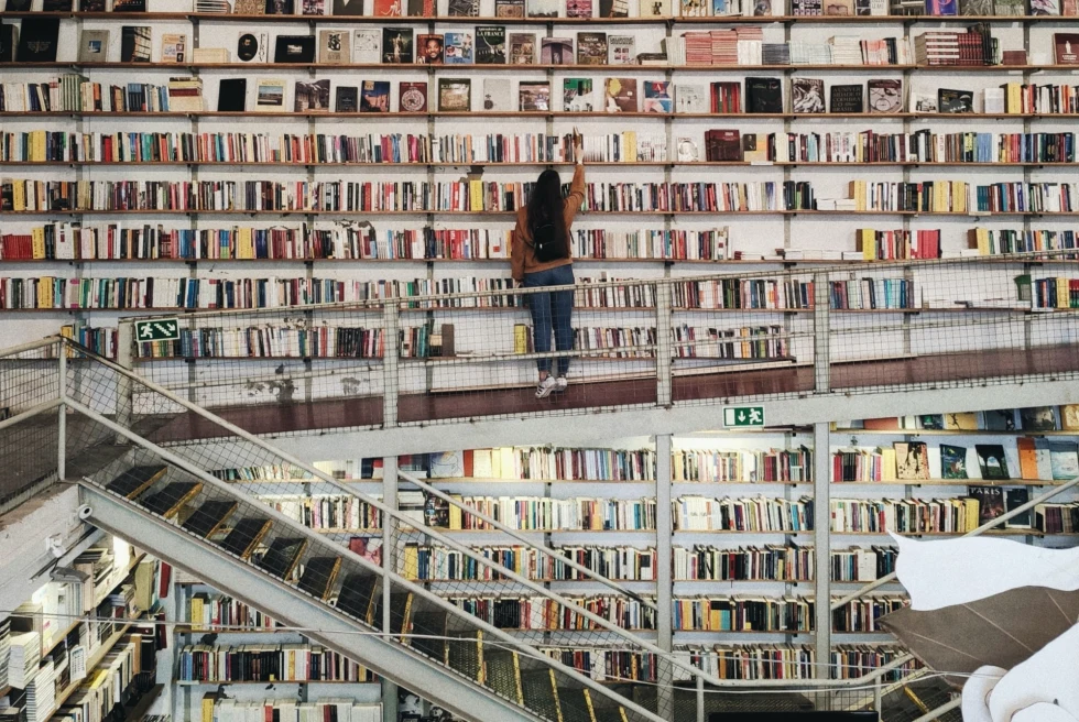 a woman picks a book in a library wall covered in books with a stairwell to a lower floor 
