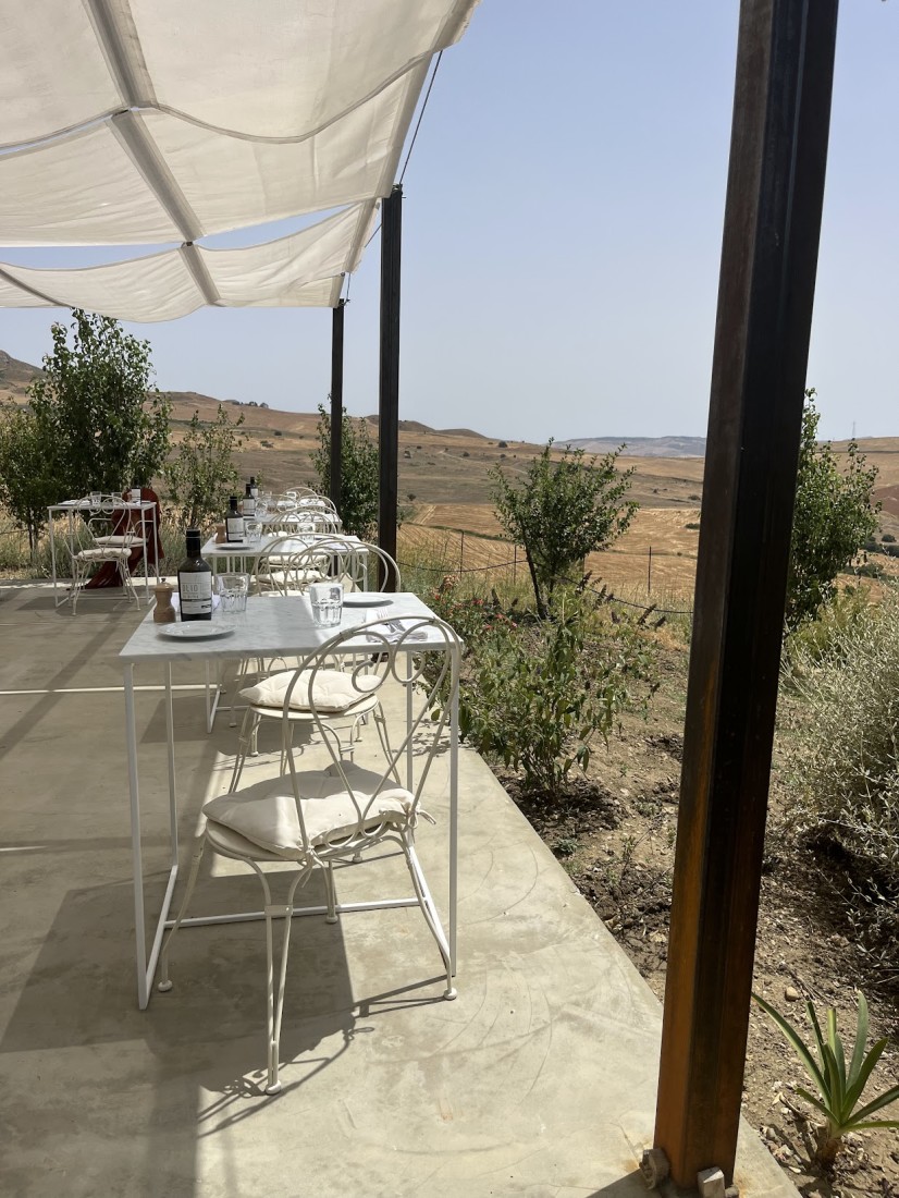 A patio with white tables and chairs, wooden beams and a white shade covering. There are bushes and desert-like terrain in the surrounding areas. 
