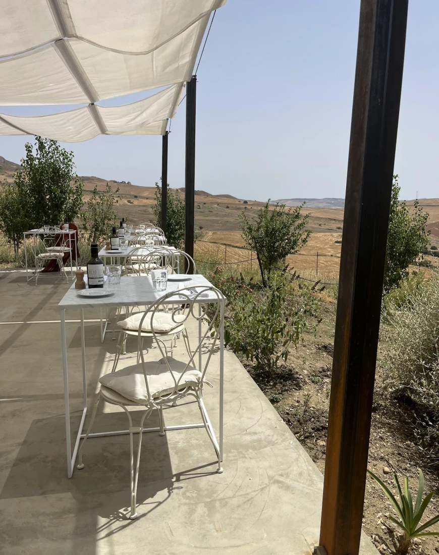 A patio with white tables and chairs, wooden beams and a white shade covering. There are bushes and desert-like terrain in the surrounding areas. 