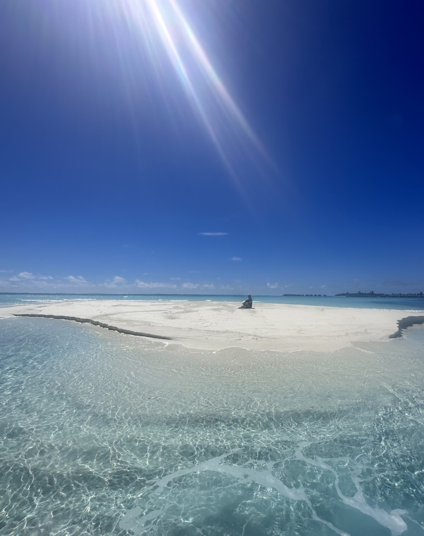 A view of the sun rays shining down onto a white sand bar surrounded by crystal clear blue water. 