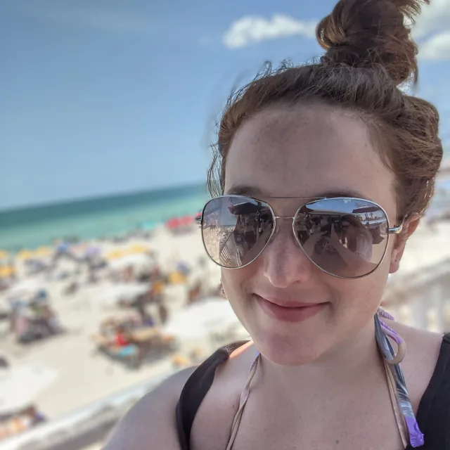 Travel Advisor Shelbie Koenitzer with large sunglasses and a black top in front of the beach.