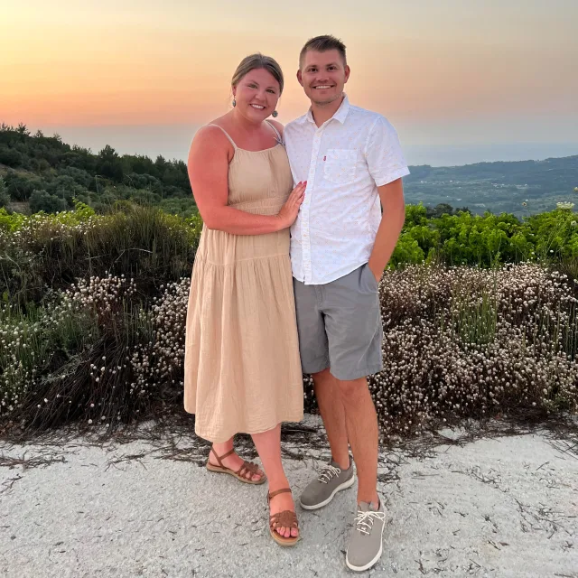 Kate Sevastakis in a beige dress posing with a man in front of a sunset. 