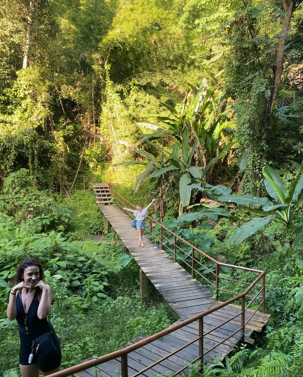 Two women posing on a path surrounded by lush and green plants and trees. 