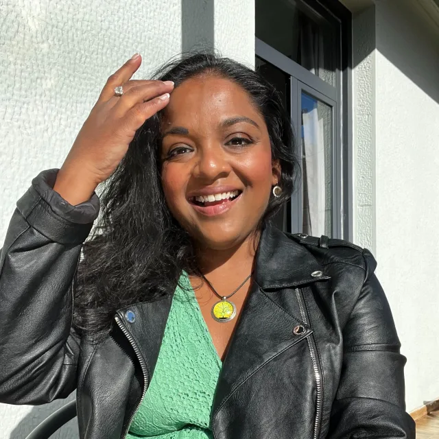 Travel Advisor Meena Thiruvengadam in a green dress and black jacket in front of a white wall.