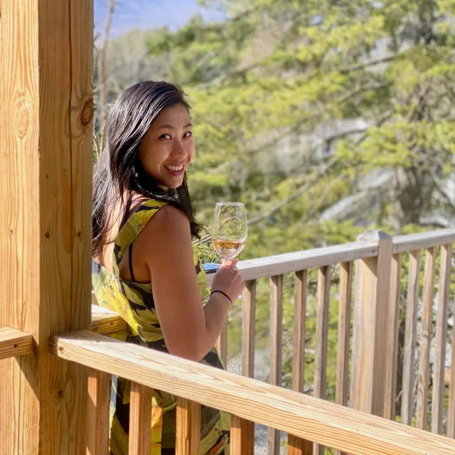 Travel Advisor Natalie Lum-Tai in a yellow and black dress, holding a wine glass in front of green trees.
