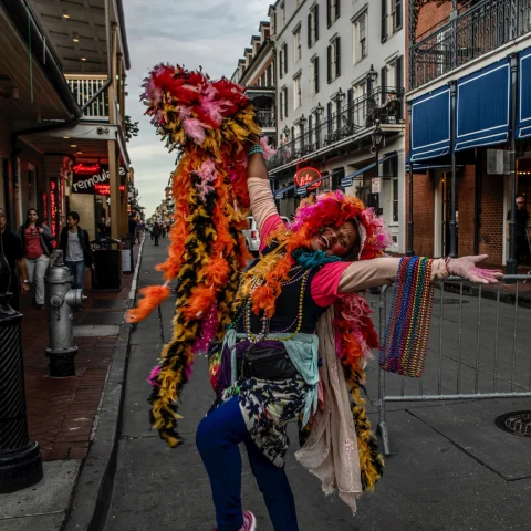 A woman adorned in flowers with her hands spread open posing in front of buildings outside in New Orleans. 