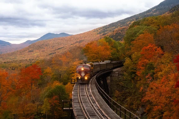 Train traveling through valley of brightly colored trees with autumns leaves on a crisp day. 