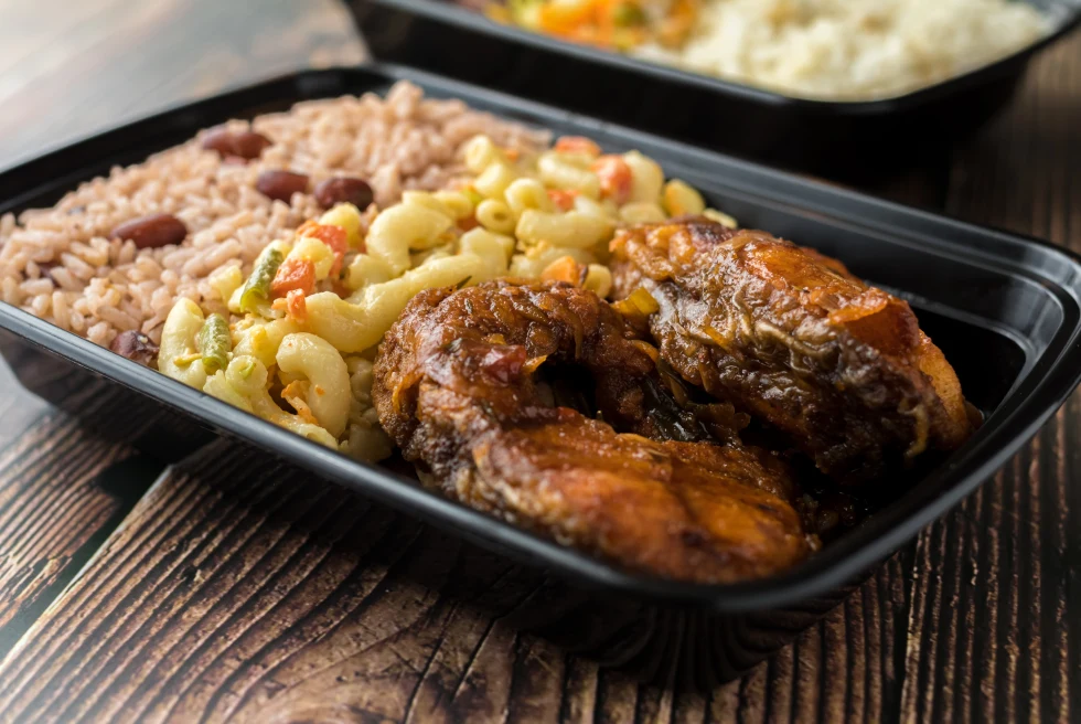 White and brown beans and rice with yellow and orange macaroni and cheese and browned chicken in a black plastic dish on a brown wooden table