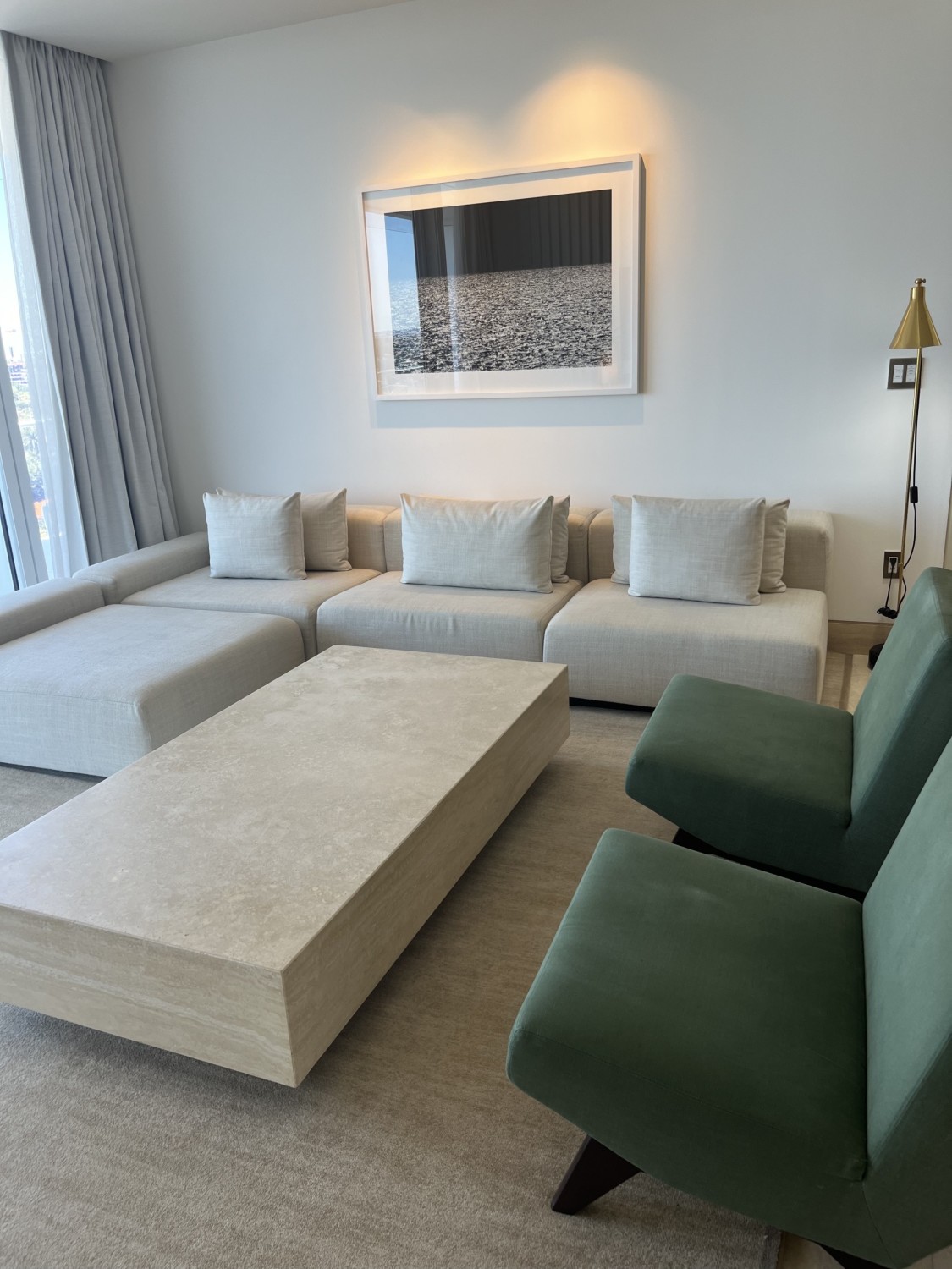 A lounge area in a hotel suite with a white couch, two green lounge chairs, artwork hanging on the white wall, a floor lamp in the right hand corner and a rectangular coffee table in the middle of the room. 