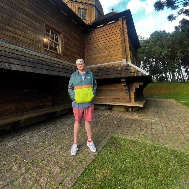 Trent Rogers posing for a photo in front of a wood cabin surrounded by trees and grass.