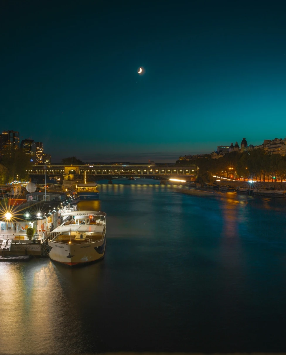 An image of a canal in the evening beneath a moon with various city lights and boats to the left of the view. 