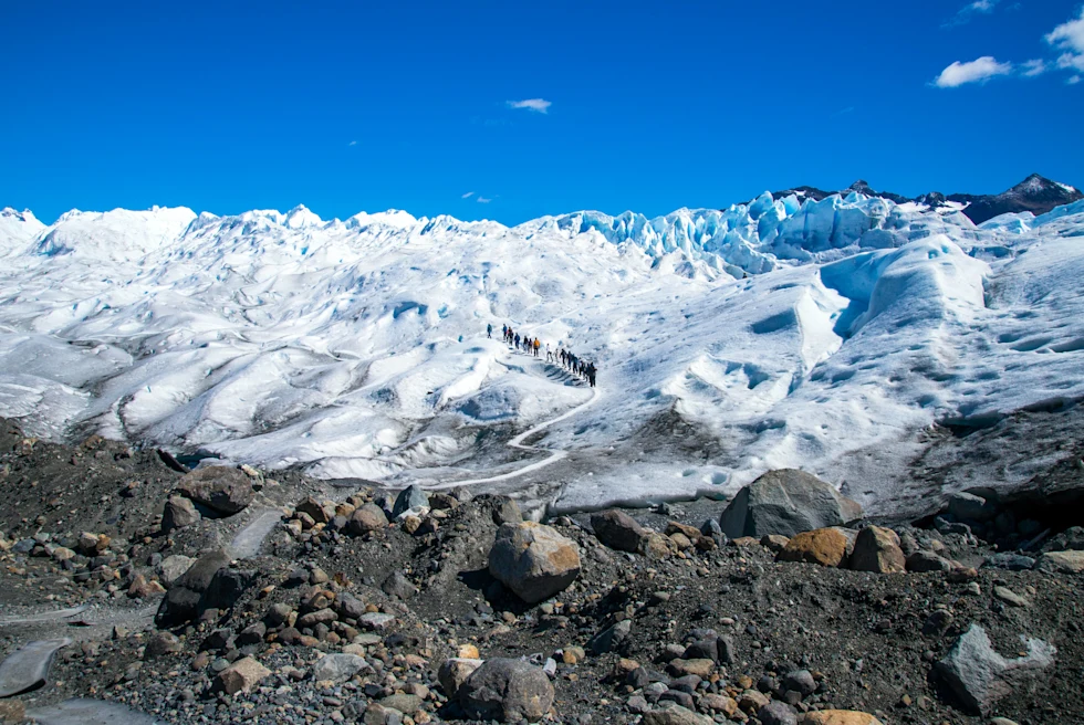 The Perito Moreno glacier in El Calafate, Argentina with brown and grey rocks at the basin of white snow with a line of people trekking in black.