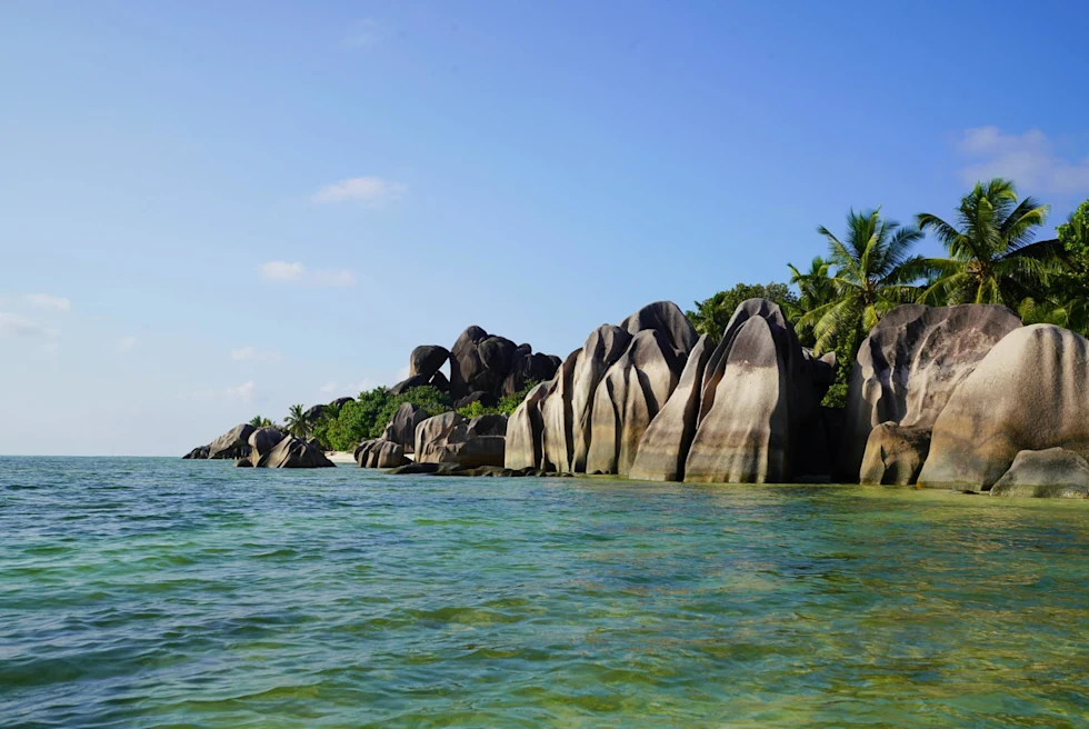 Turquoise waters in La Digue against gray and brown striped rocks and palm trees.