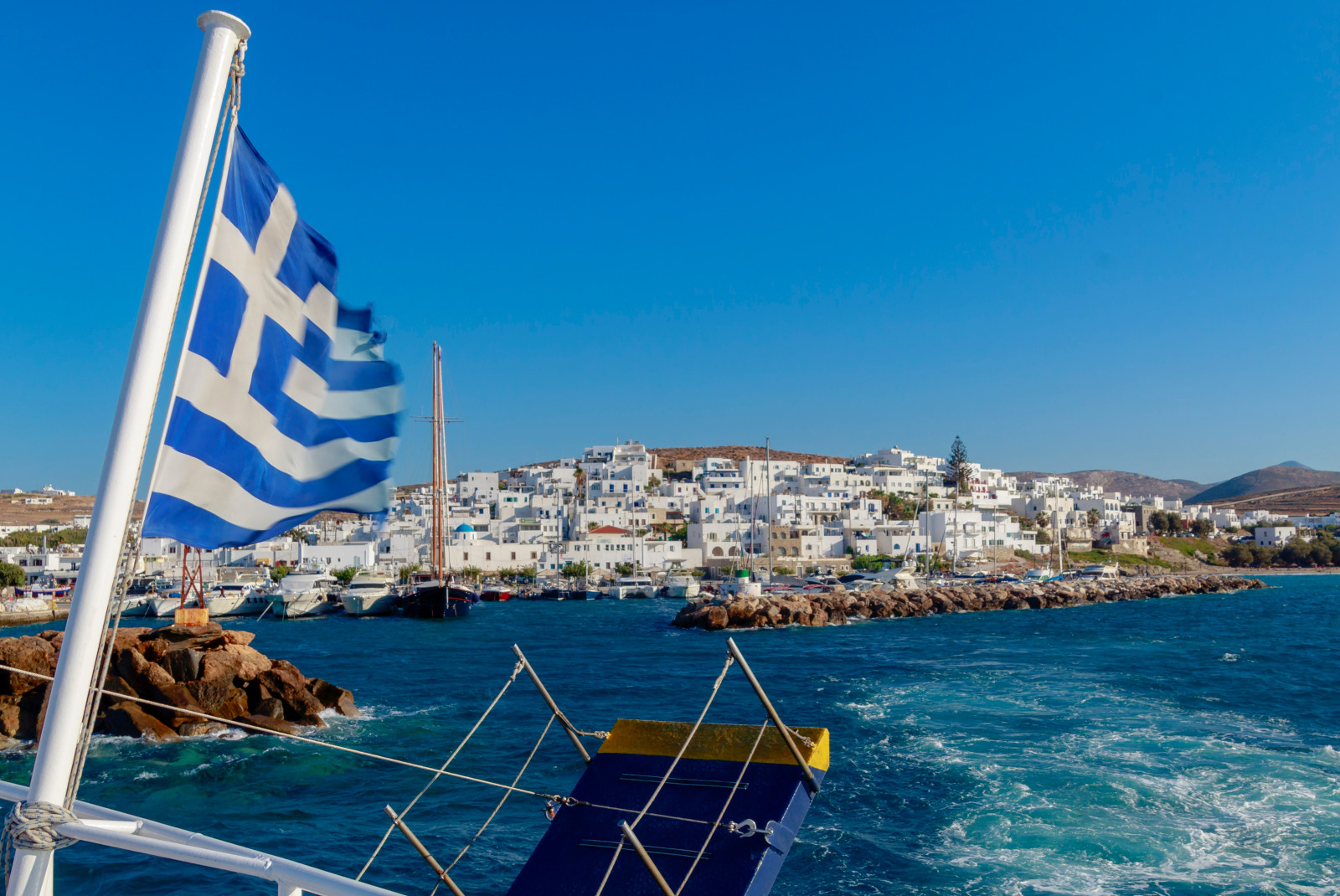 Body of water next to blue and white flag waving with white buildings in the distance during daytime