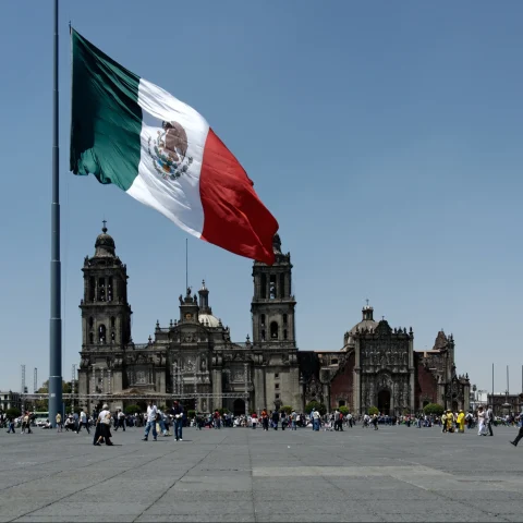 A view of the flag in Mexico in front of people walking near a grand stone building. 